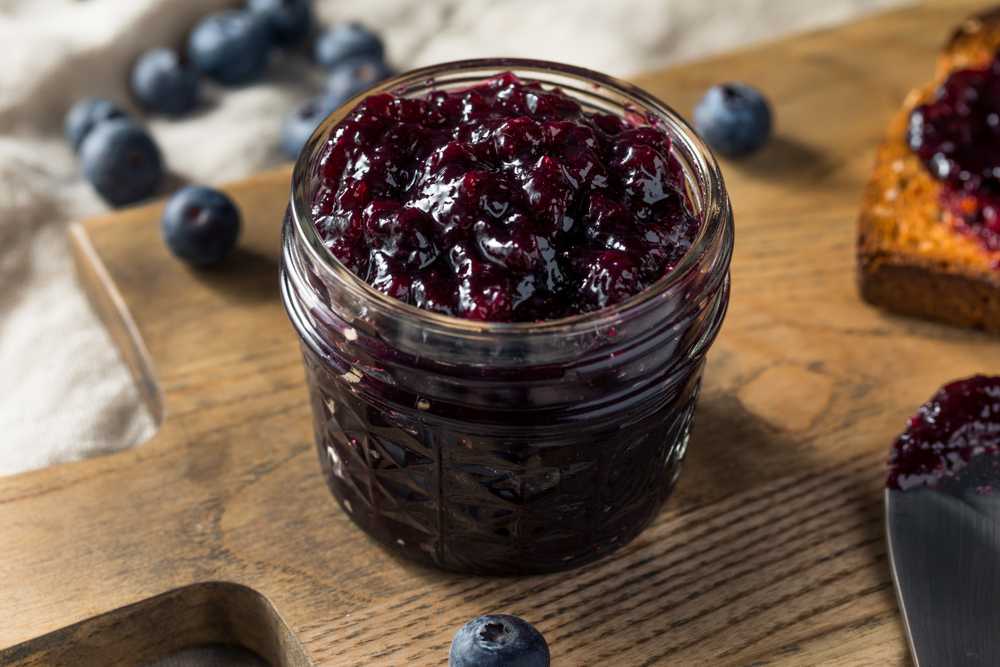 A glass jar of blueberry jam on a wooden board, with fresh blueberries scattered nearby.