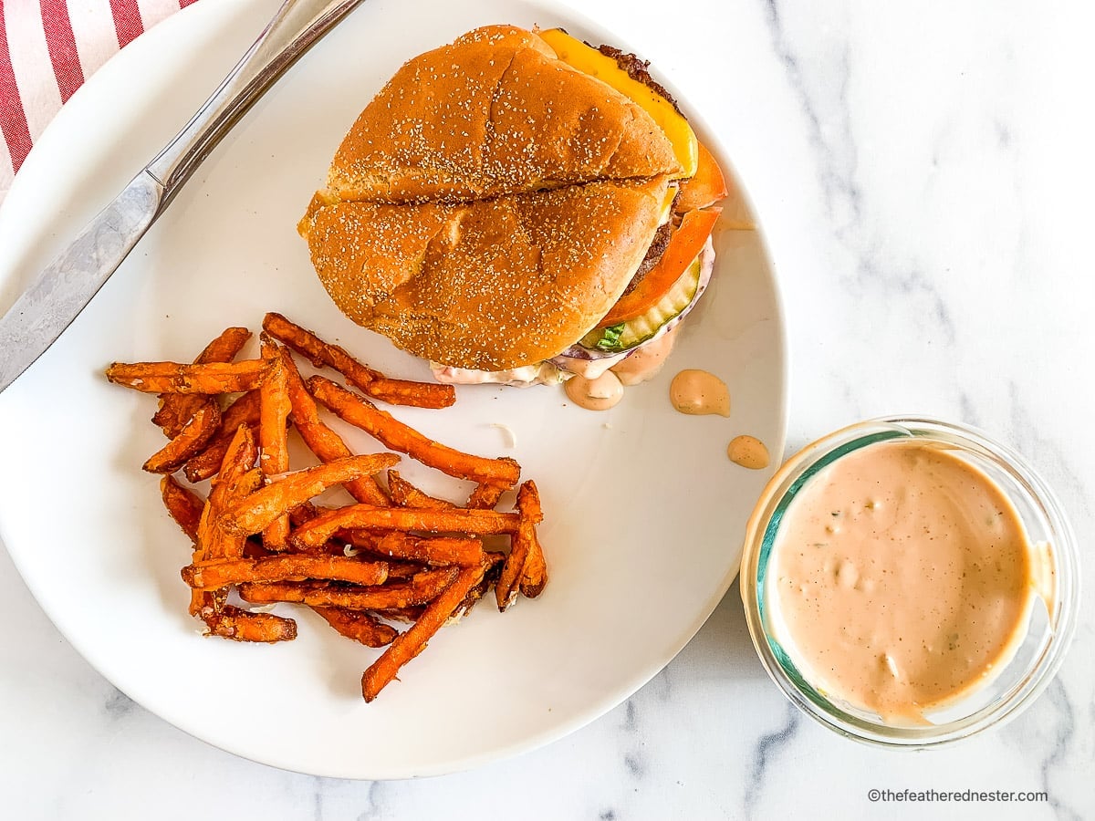 A burger with sweet potato fries and dipping sauce on a white plate, next to a knife and red-striped napkin.