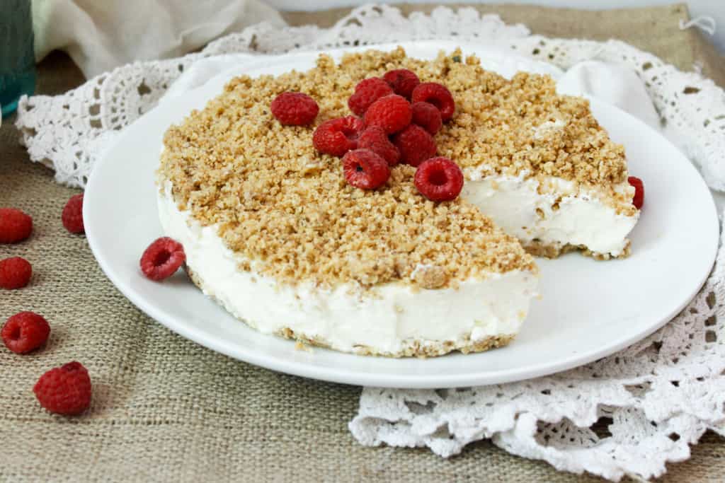 No-bake cheesecake topped with crumbled crust and fresh raspberries on a white plate, with raspberries scattered nearby.