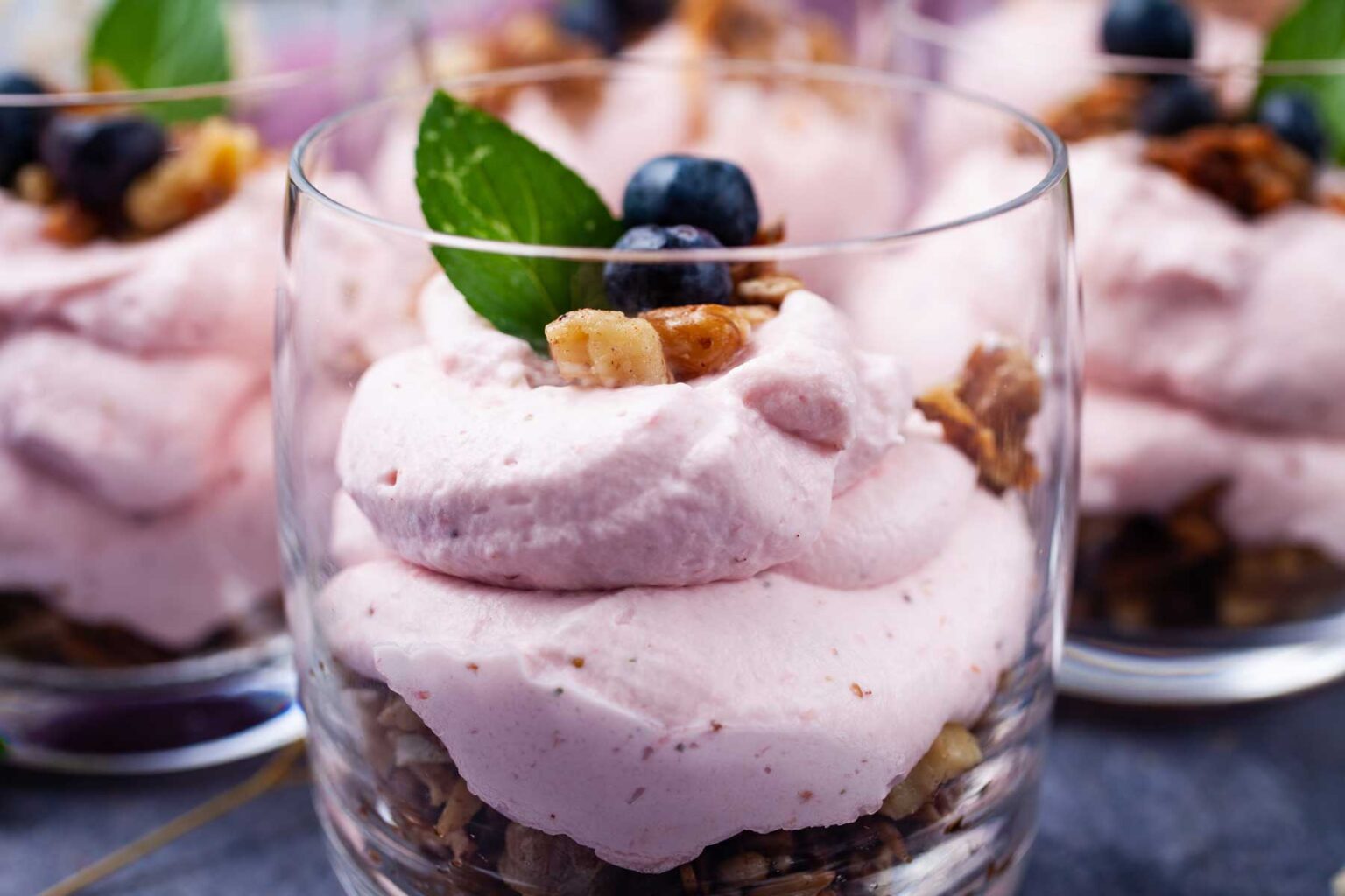 A dessert glass with pink mousse, granola, blueberries, and a mint leaf on top.