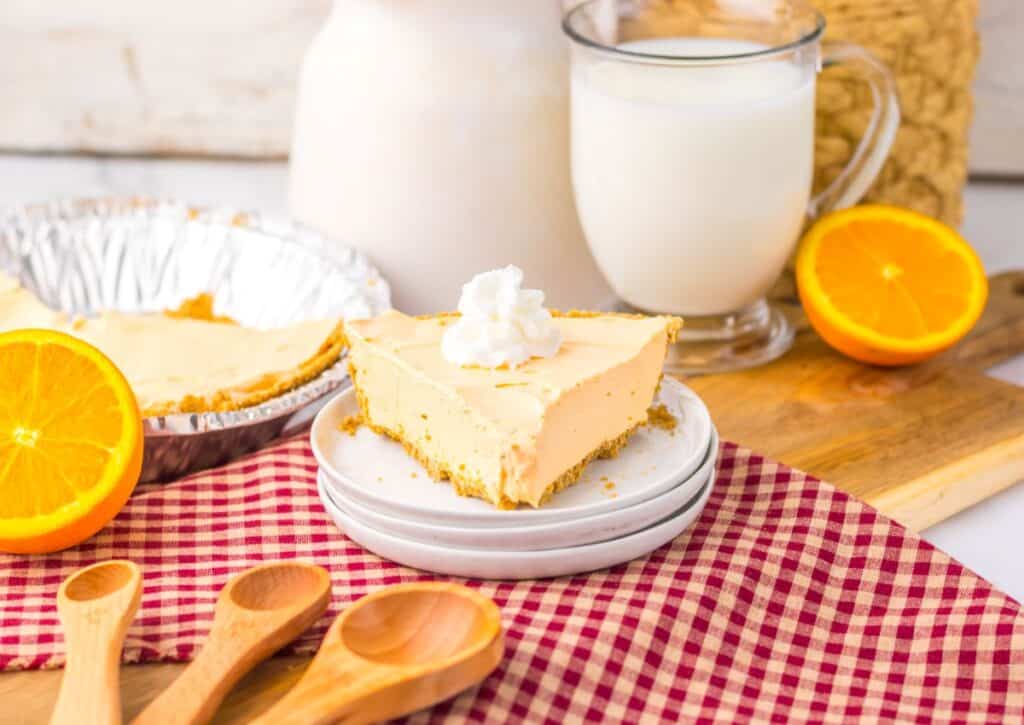 A slice of creamy pie with whipped topping, orange halves, and a glass of milk on a red checked cloth.