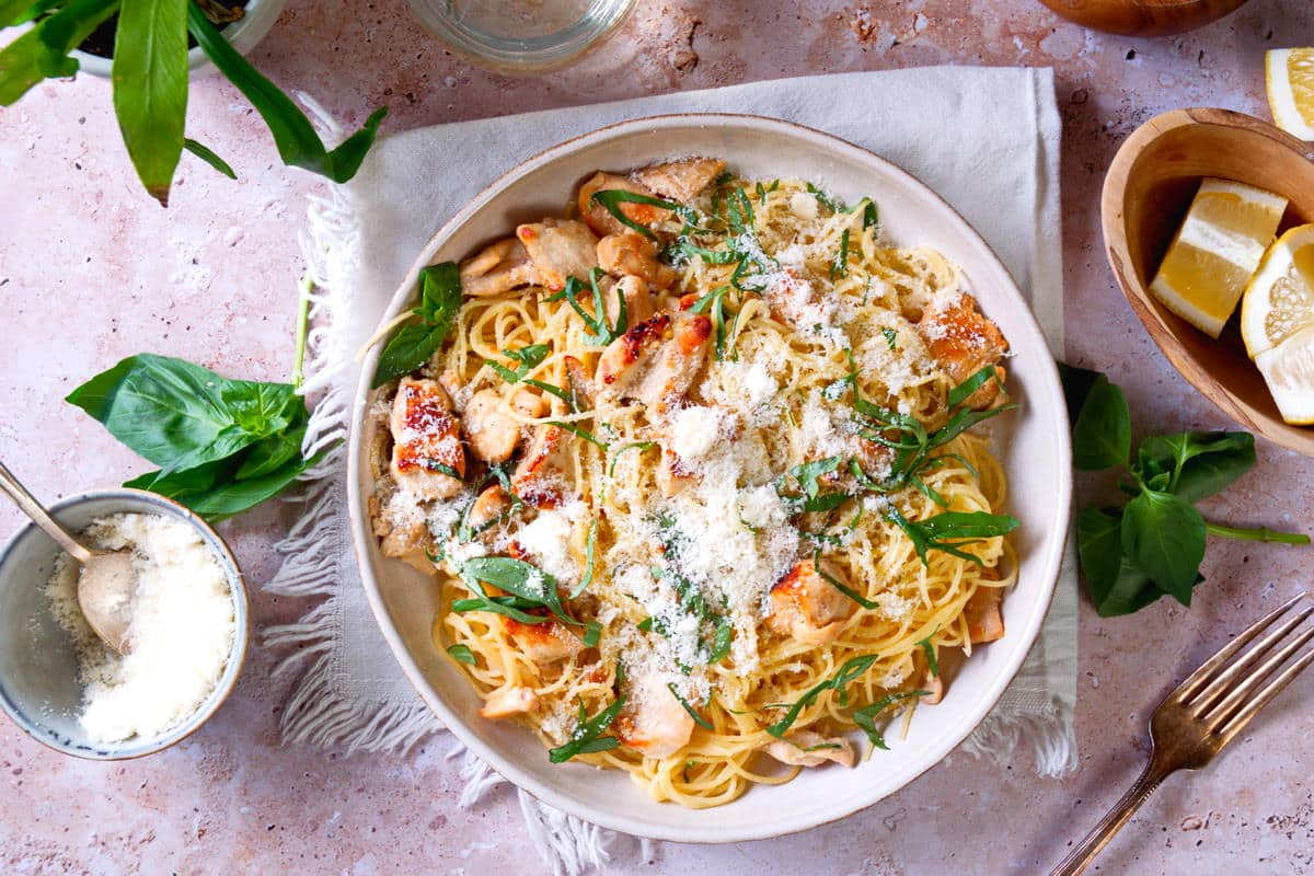 A plate of creamy pasta with grilled chicken, grated cheese, and fresh green herbs on top.