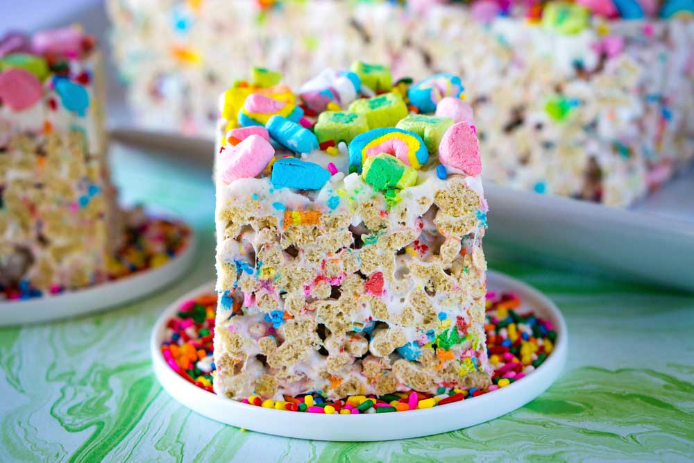 A colorful marshmallow cereal treat topped with Lucky Charms and sprinkles on a white plate.