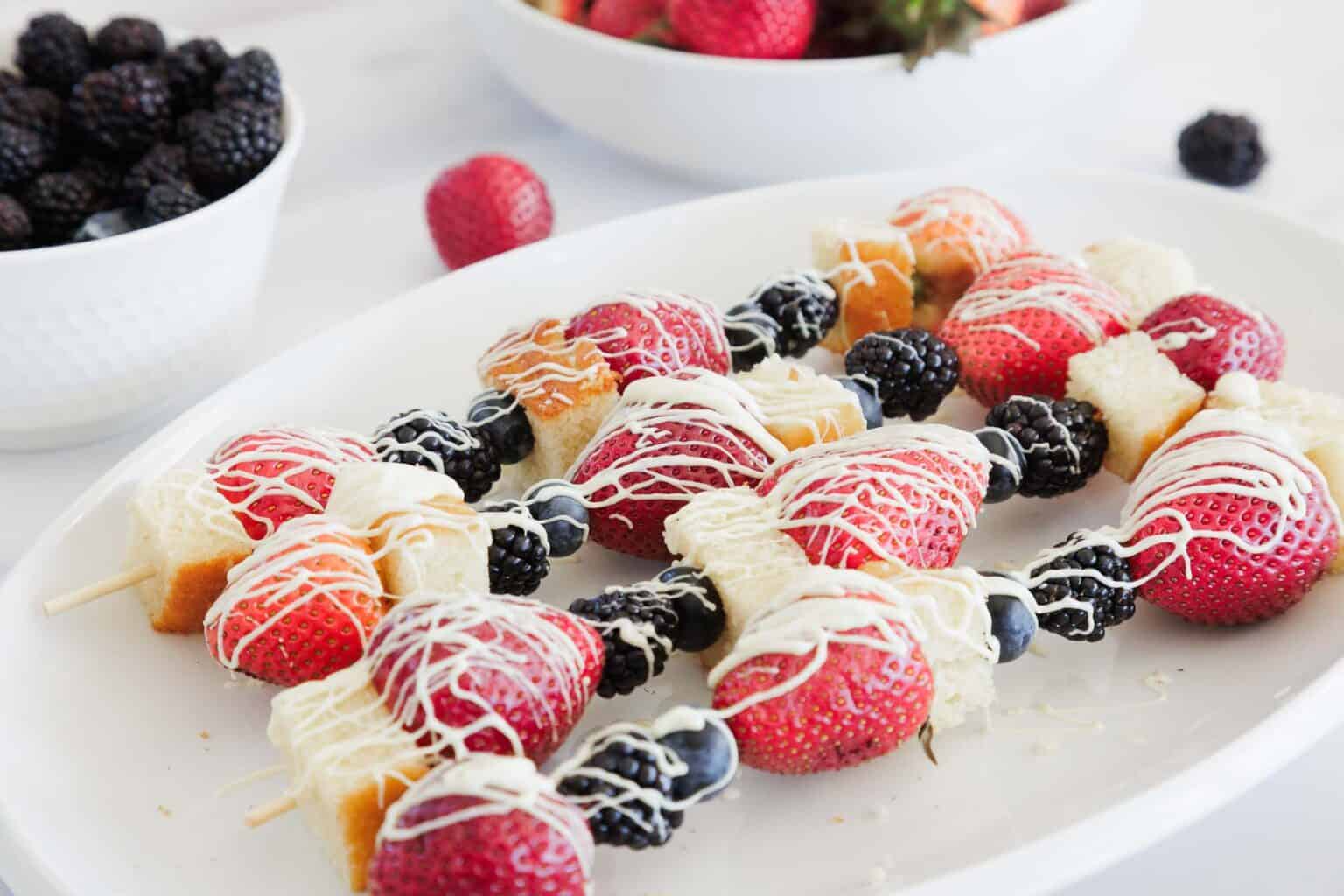 Fruit and cake skewers drizzled with white chocolate on a white plate, with berries in the background.