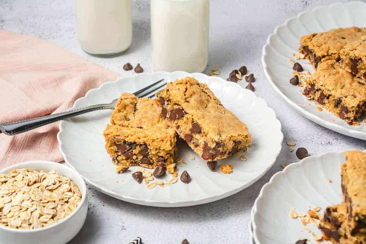 Two chocolate chip oatmeal bars on a white plate with a fork, oats, and glasses of milk in the background.