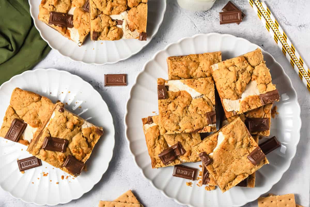 Plates of s’mores cookie bars with Hershey’s chocolate pieces and graham crackers on a white surface.