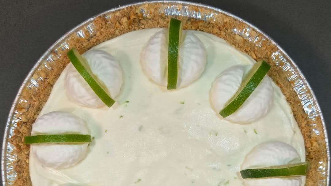A key lime pie with whipped cream dollops and lime slices on a graham cracker crust in a foil pie tin.