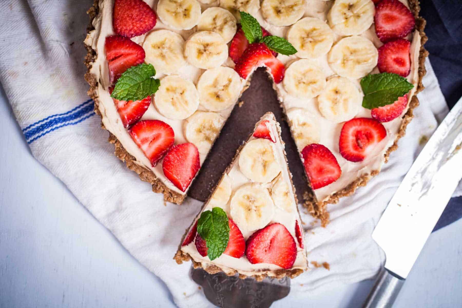 A fruit tart topped with sliced bananas, strawberries, and mint, with one slice cut and placed in front.