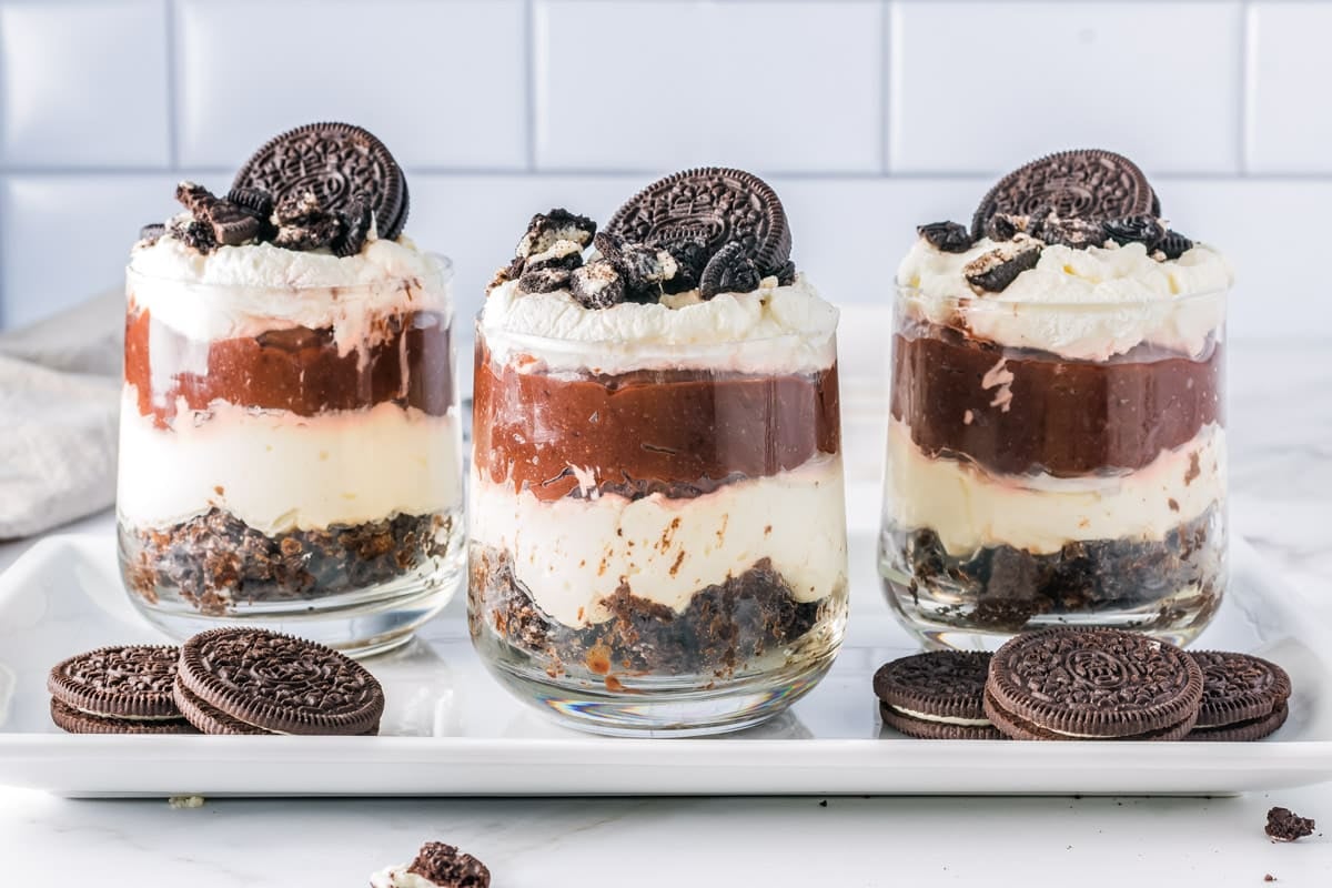 Three layered dessert parfaits with whipped cream and chocolate cookies in glass cups, topped with whole cookies.