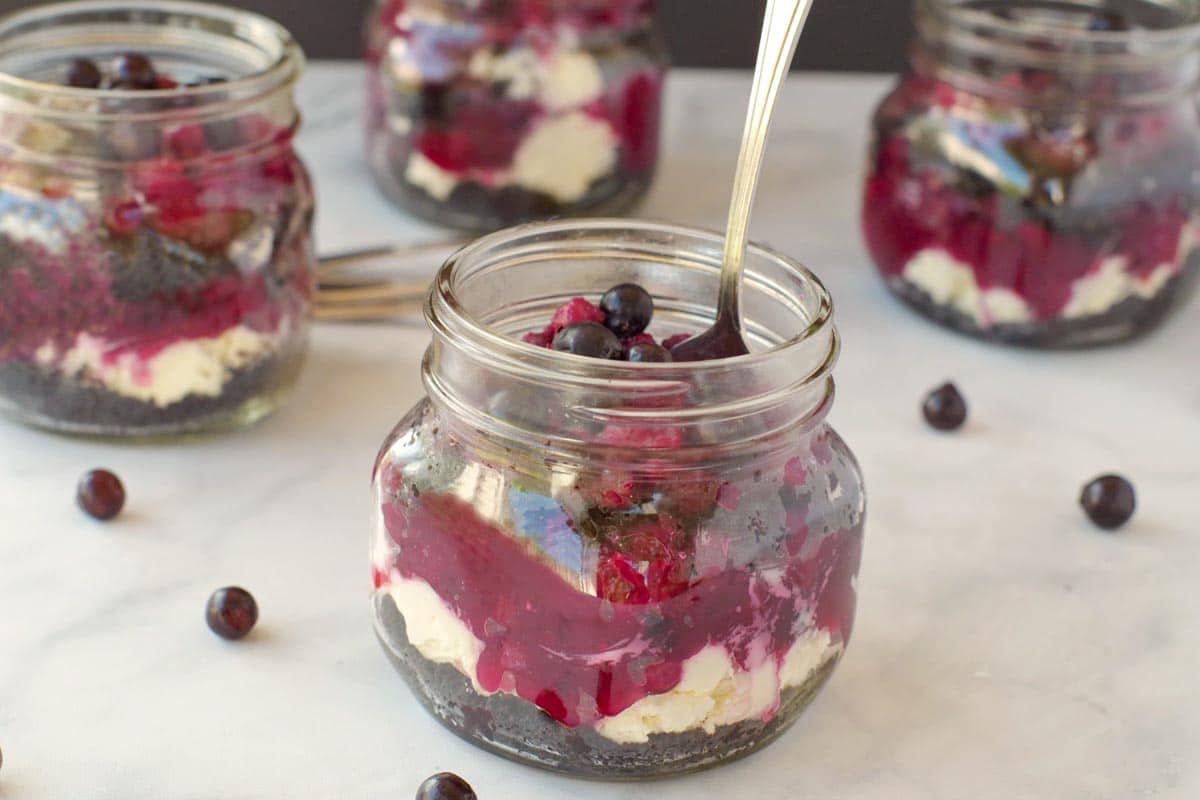 A mason jar dessert with layers of cream, berry sauce, and crust, topped with blueberries on a marble surface.