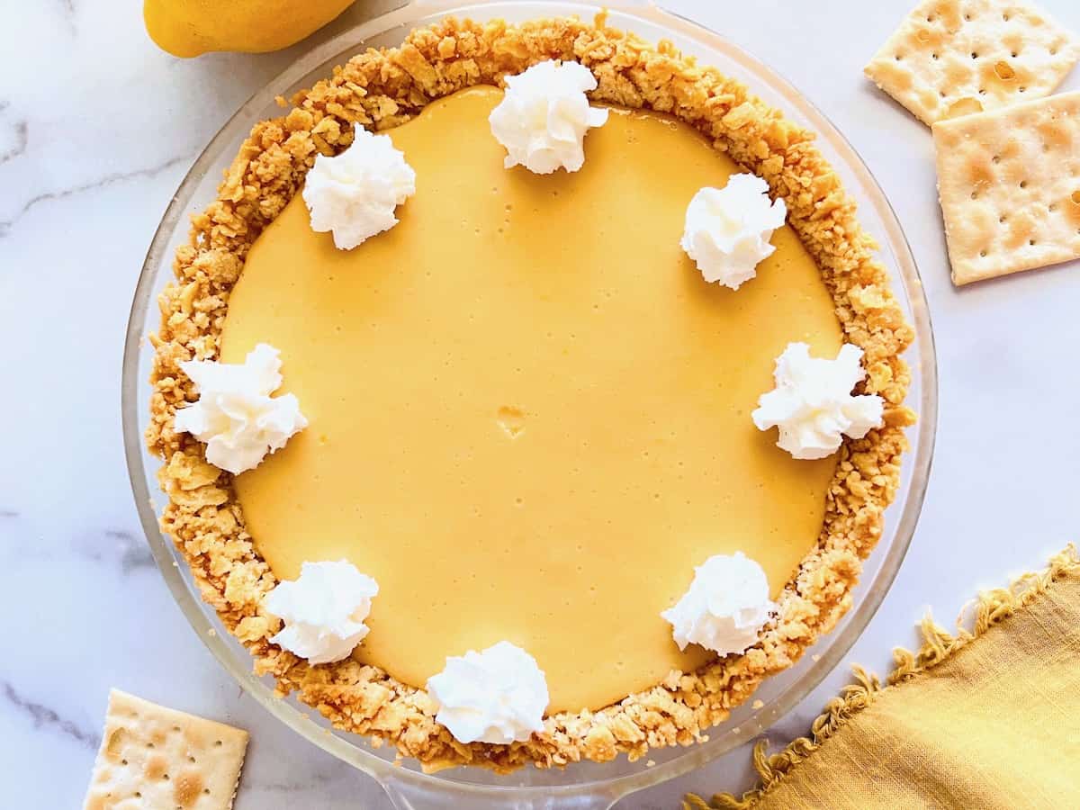 Lemon pie with whipped cream dollops on top, surrounded by crackers on a marble surface.
