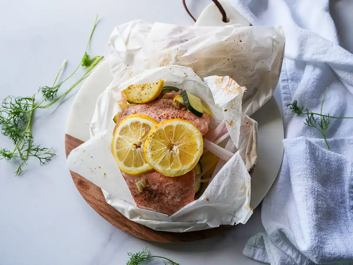 Salmon fillet baked in parchment paper with lemon slices and herbs on a white plate.