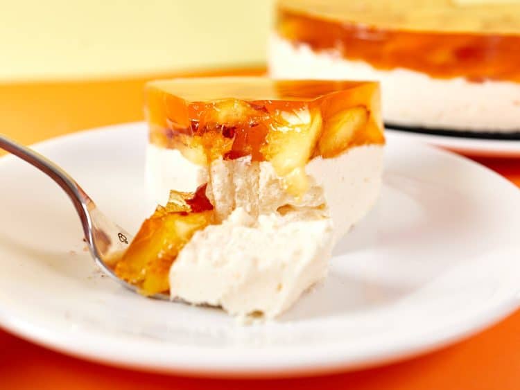 A forkful of creamy cheesecake with fruit jelly topping on a white plate, with the whole cake in the background.