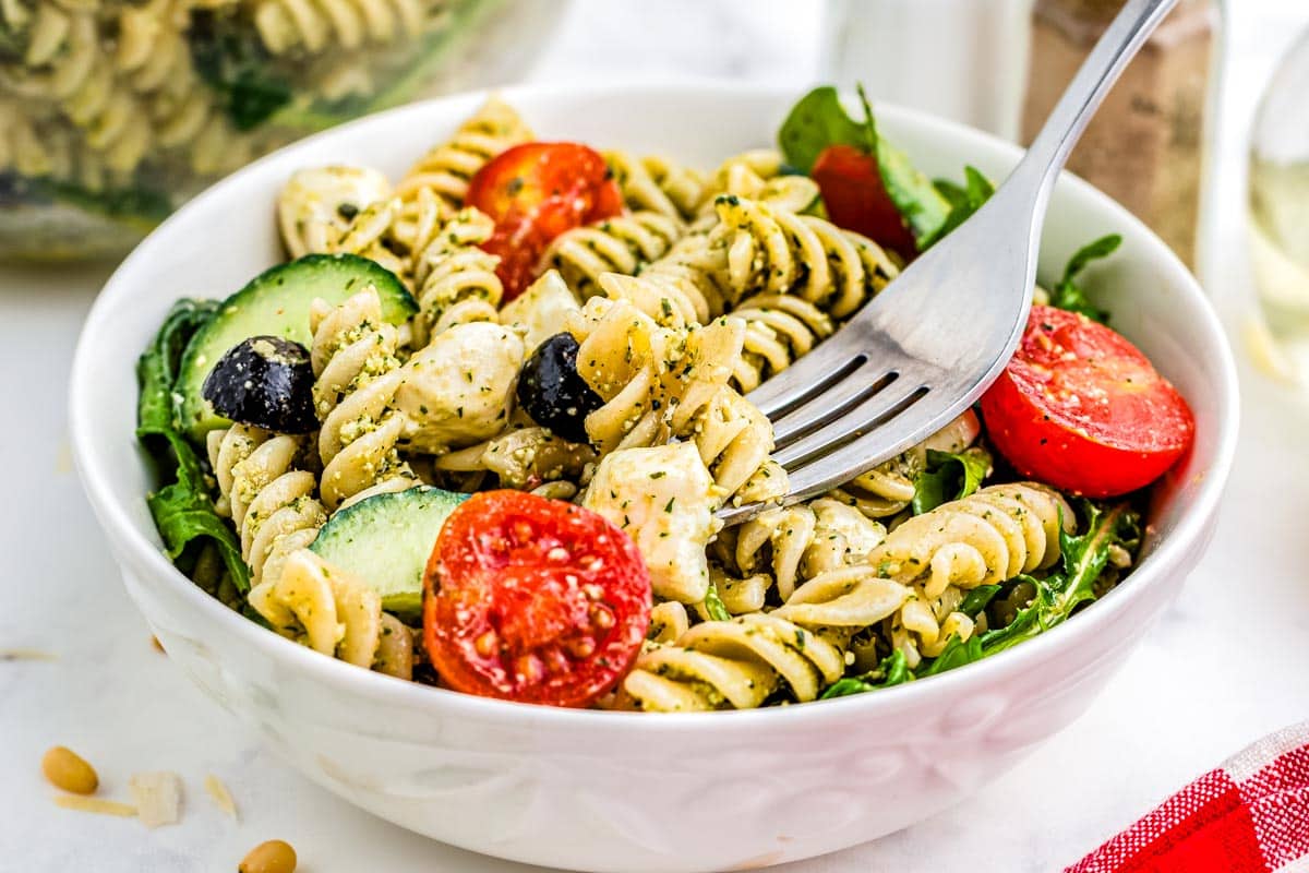 Pasta salad with rotini, cherry tomatoes, cucumber, olives, mozzarella, and greens in a white bowl with a fork.