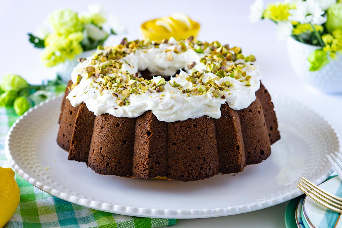 Bundt cake topped with whipped cream and chopped pistachios, displayed on a white plate with flowers nearby.