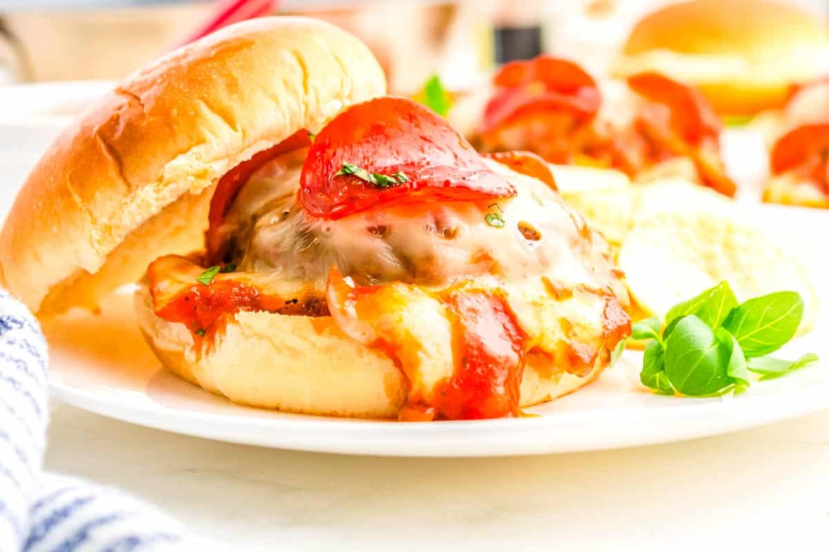 A hamburger topped with melted cheese, pepperoni, and tomato sauce, served on a white plate with chips.