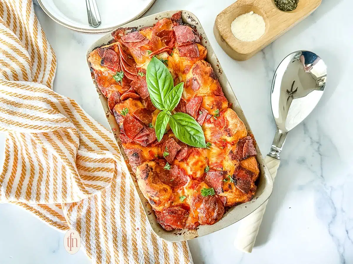 A baked pepperoni pizza casserole garnished with basil leaves, next to a striped towel and a serving spoon.