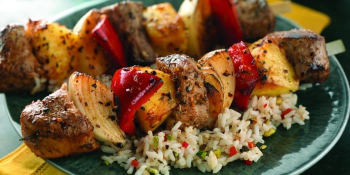 Grilled beef and vegetable skewers served over rice with red peppers, onions, and pineapple on a green plate.