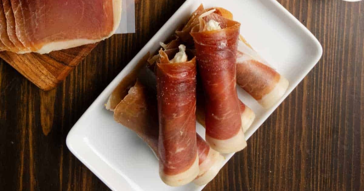 Three slices of prosciutto rolled and served on a white rectangular plate on a wooden table.