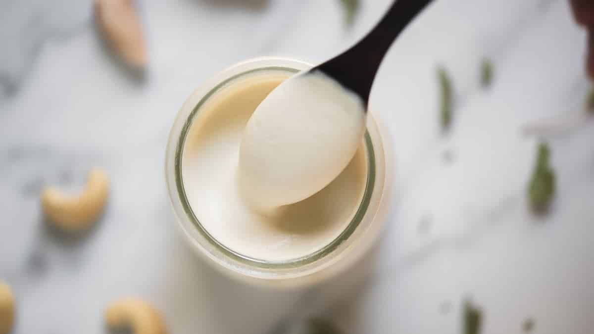 A spoon scooping creamy white yogurt from a glass jar on a marble surface with scattered cashews.