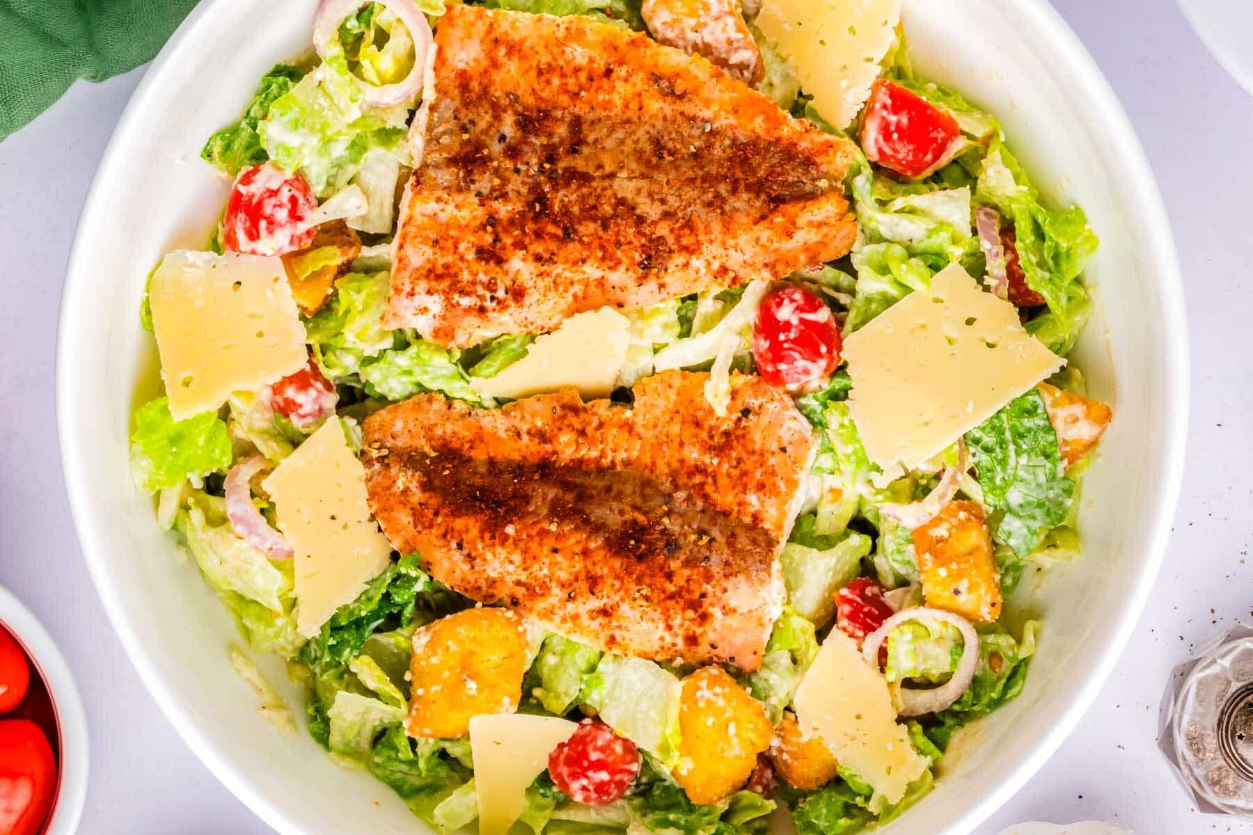 Caesar salad topped with two pieces of seasoned grilled salmon, cherry tomatoes, croutons, and shaved parmesan.