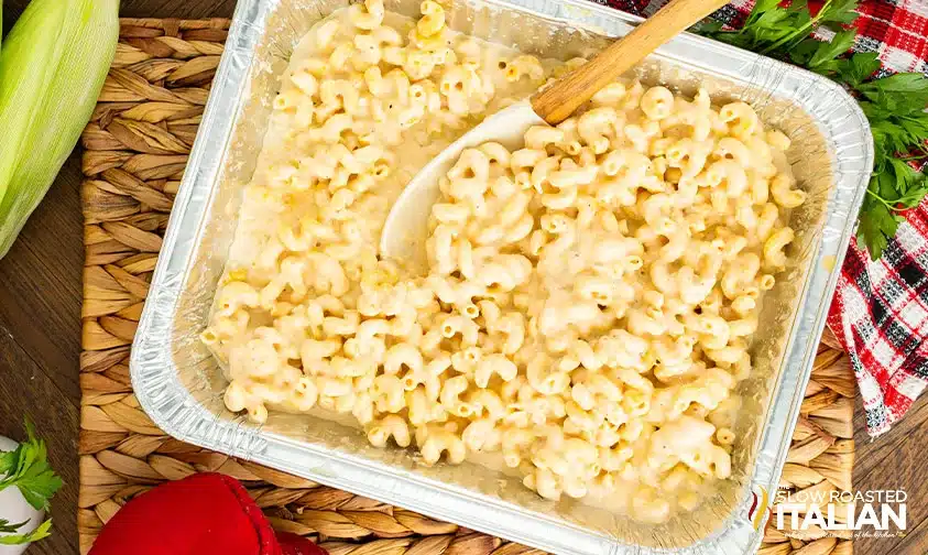 A tray of creamy macaroni and cheese with a serving spoon, on a wooden surface next to herbs and a red plaid cloth.