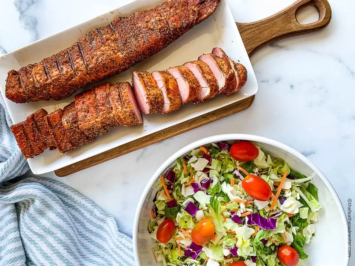 Sliced seasoned pork tenderloin on a tray next to a bowl of colorful salad with tomatoes and cabbage.