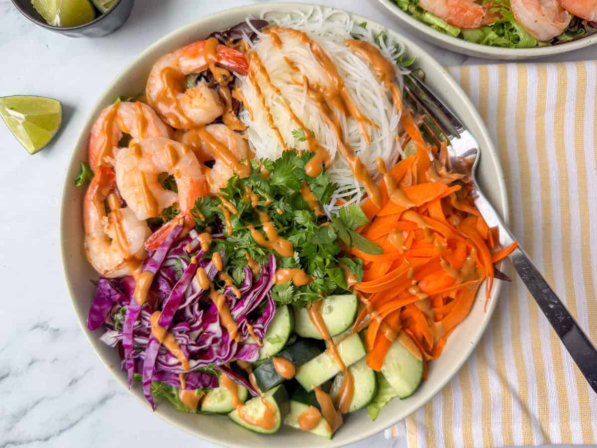 A bowl of noodles topped with shrimp, cucumber, carrots, purple cabbage, cilantro, and a creamy sauce.