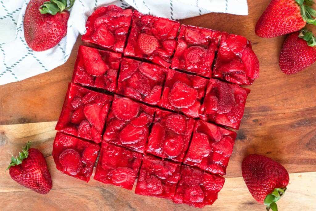 Sixteen strawberry-topped dessert bars are arranged on a wooden board, surrounded by fresh strawberries.