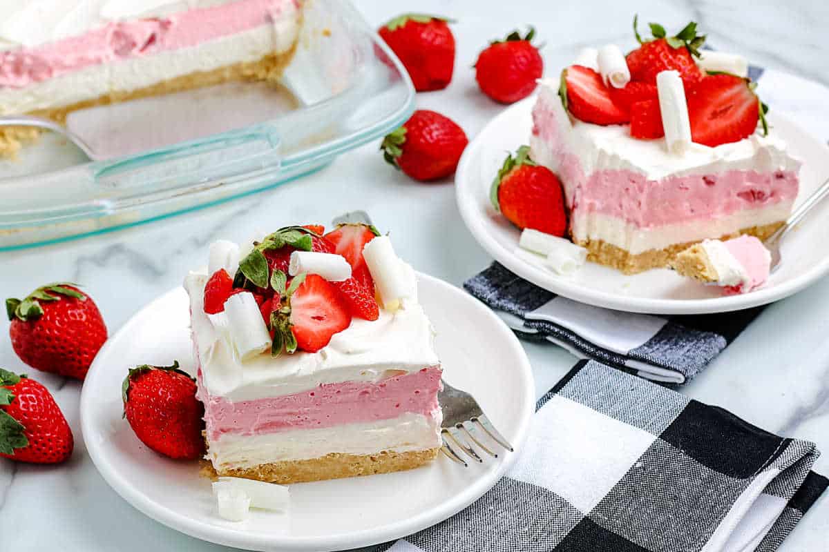 Two plates with layered strawberry dessert topped with strawberries and whipped cream, next to a baking dish.