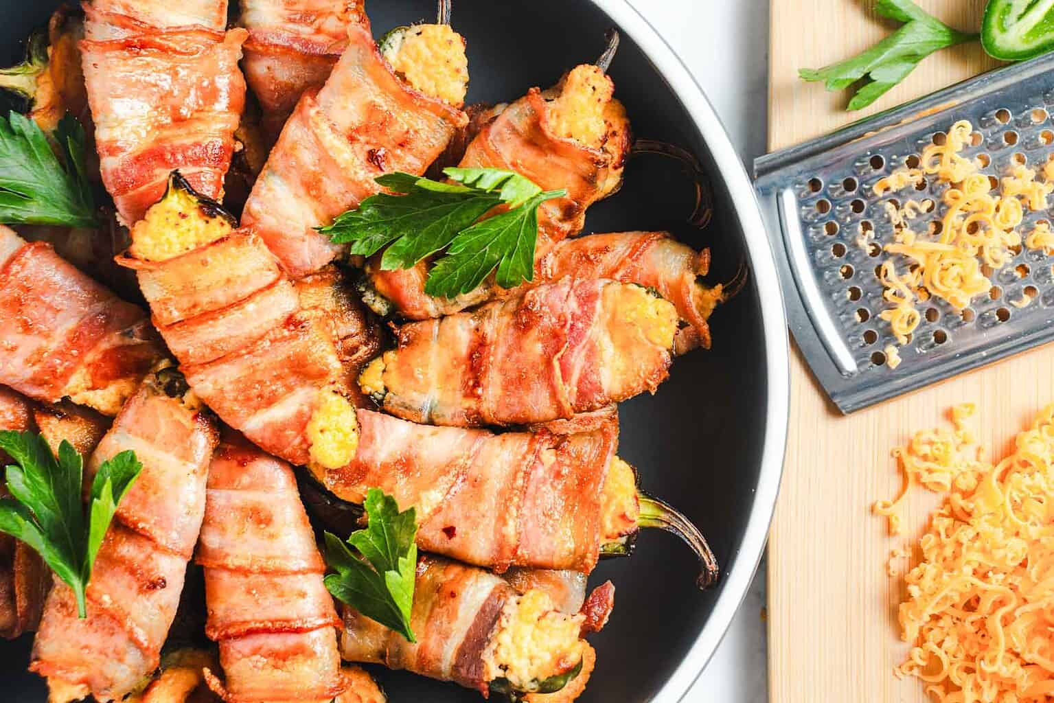 Bacon-wrapped stuffed jalapeños in a bowl, garnished with parsley, beside shredded cheese and a grater.