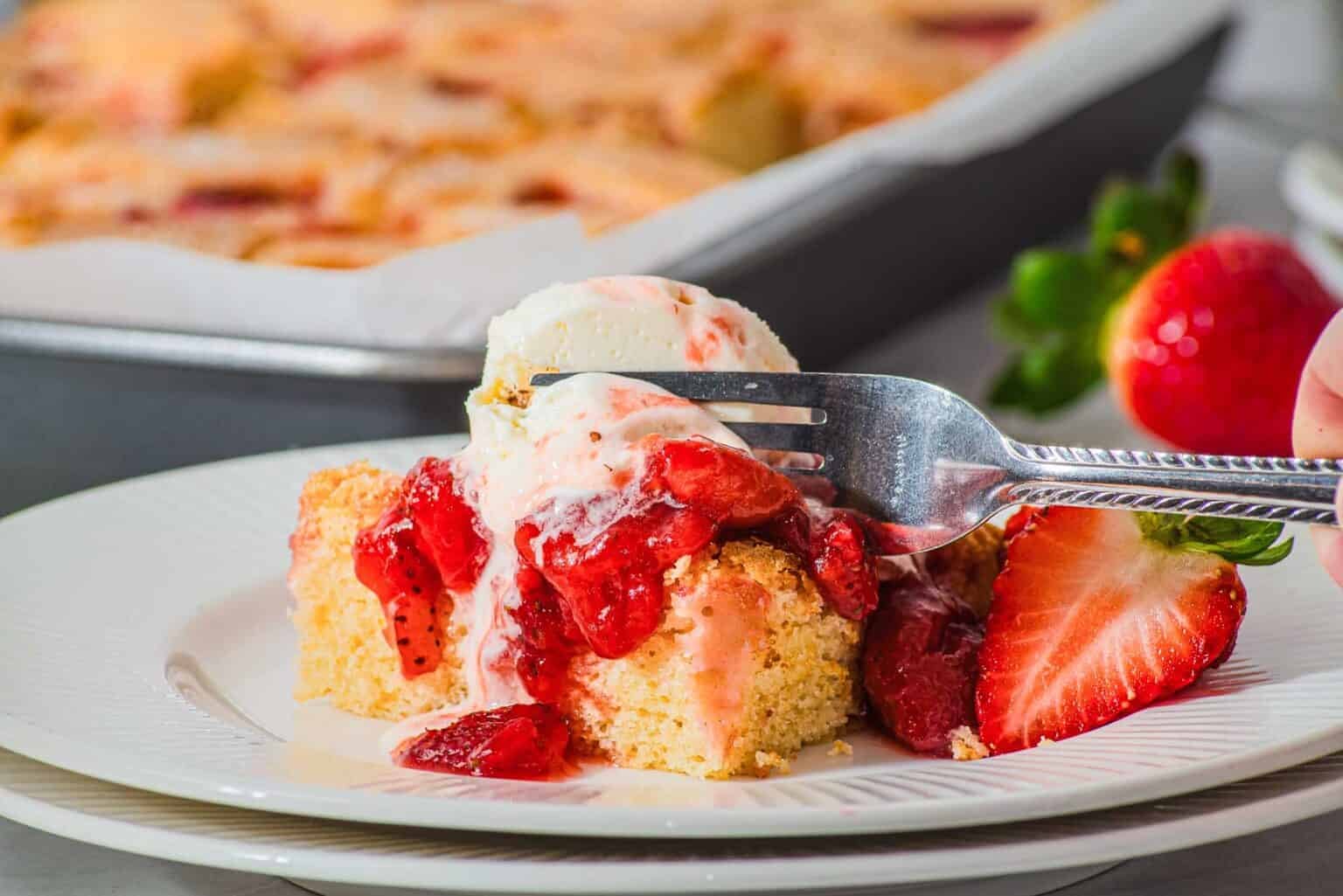 A fork cuts into strawberry shortcake topped with ice cream and fresh strawberries on a white plate.
