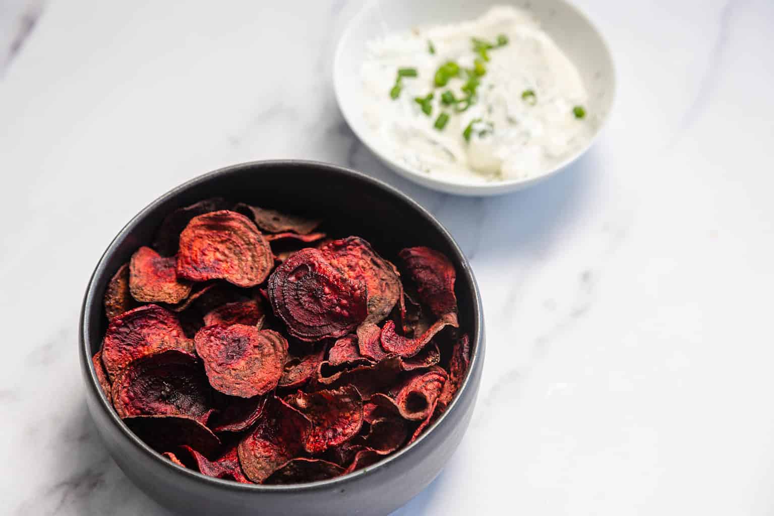 A bowl of beet chips next to a bowl of dip topped with chopped chives on a white surface.