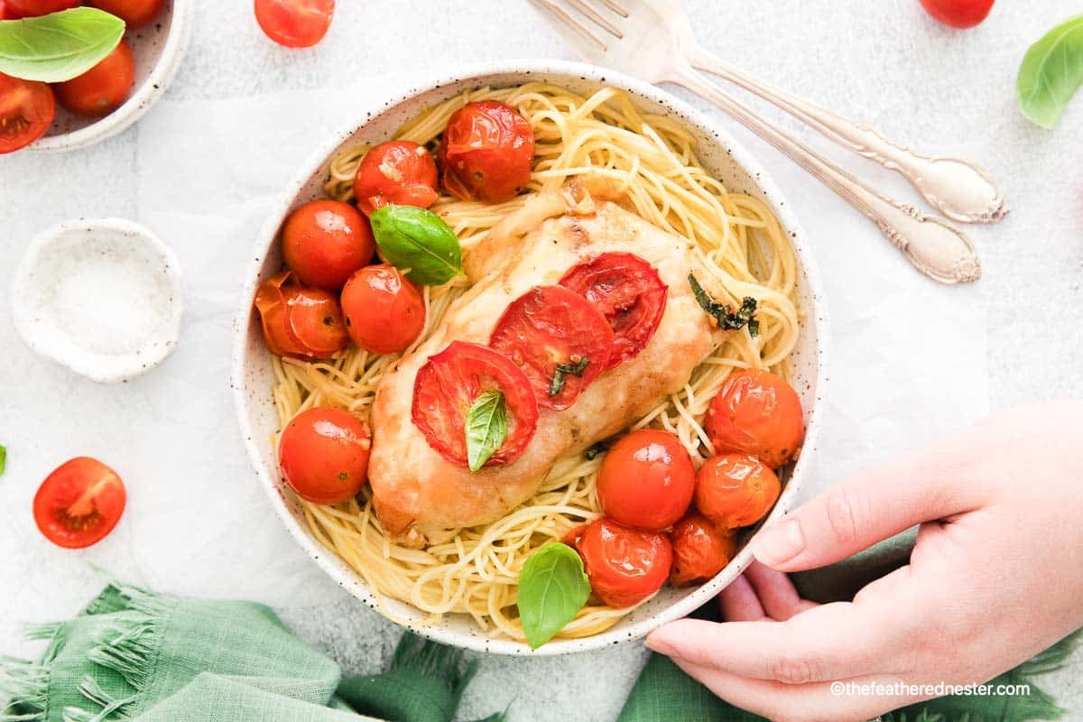 Bowl of spaghetti topped with baked chicken, cherry tomatoes, basil, and two slices of tomato, with a hand holding it.