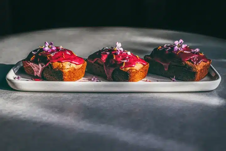 Three small cakes with pink icing and purple flowers on top, arranged on a rectangular white plate.