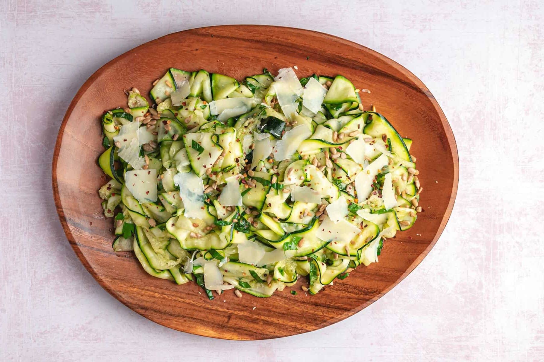 Thinly sliced zucchini salad topped with shaved cheese and seeds on a wooden plate.