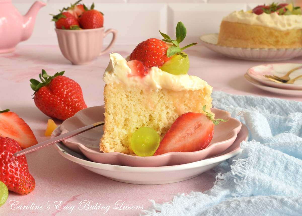 A slice of sponge cake with cream, strawberries, and grapes on a pink plate, with whole cakes and fruit nearby.