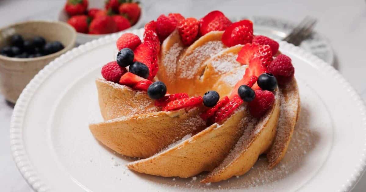 A spiral bundt cake topped with strawberries, blueberries, and powdered sugar on a white plate.