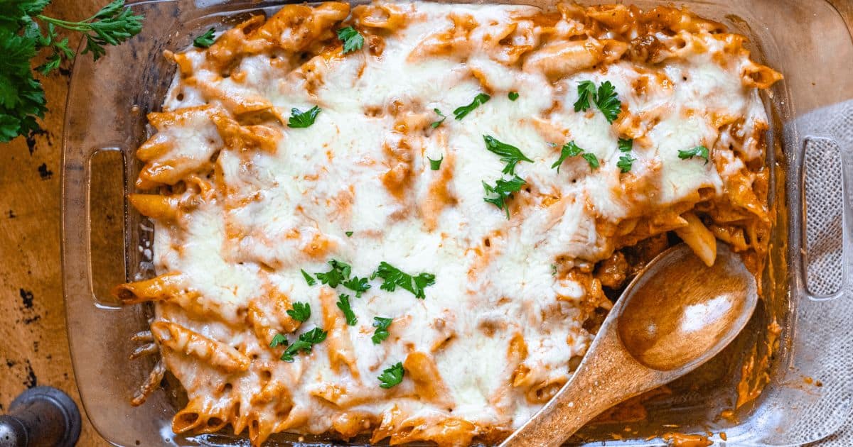 Baked pasta with melted cheese, tomato sauce, and parsley in a glass dish with a wooden spoon.