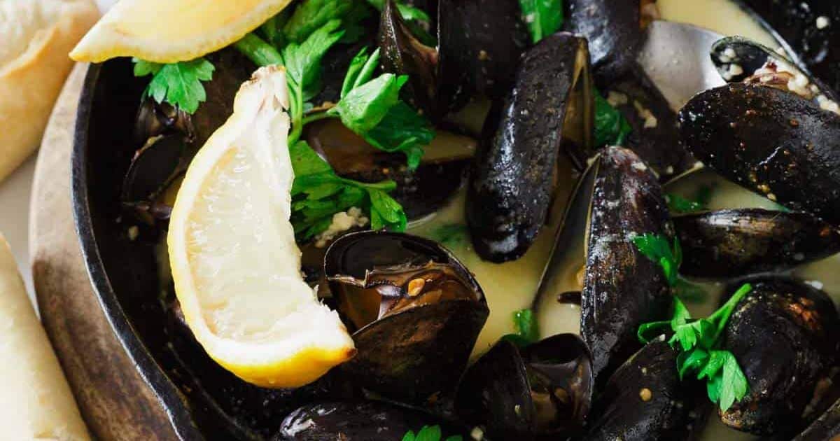 Close-up of cooked mussels in broth, garnished with parsley and lemon wedges.