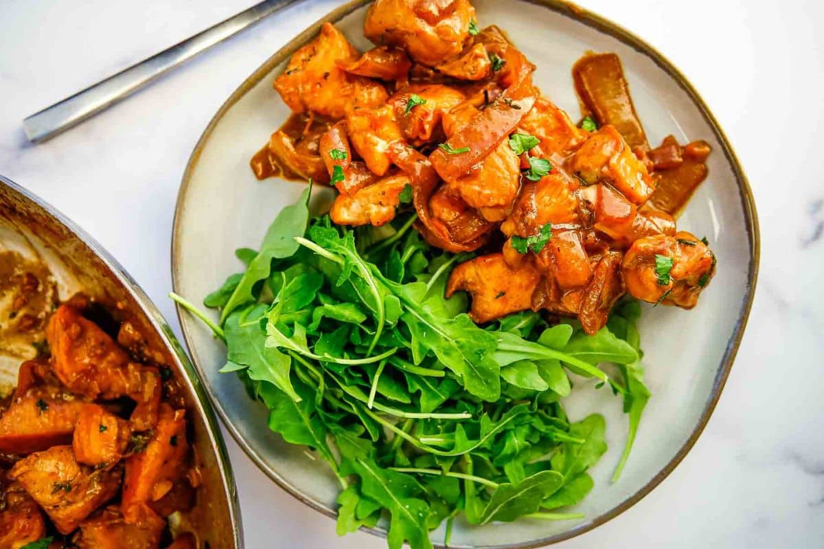 Plate of saucy chicken with onions and herbs, served next to a fresh arugula salad on a white plate.