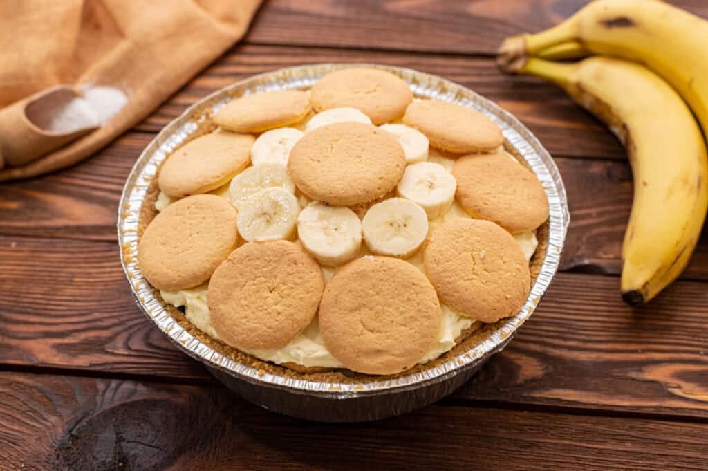 A banana pudding pie topped with vanilla wafers and banana slices, with bananas and a napkin in the background.