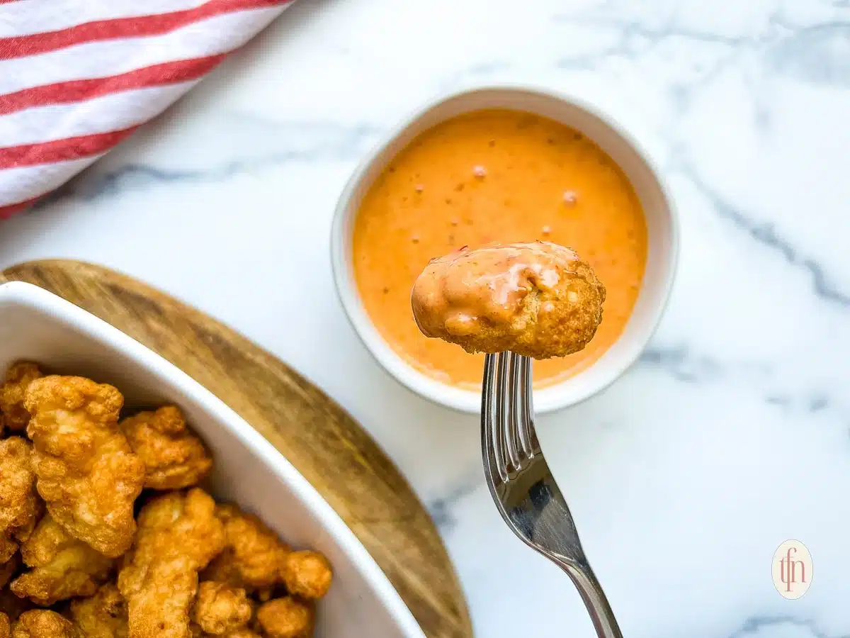 A fork holds a chicken nugget dipped in orange sauce, with more nuggets and sauce nearby on a white surface.