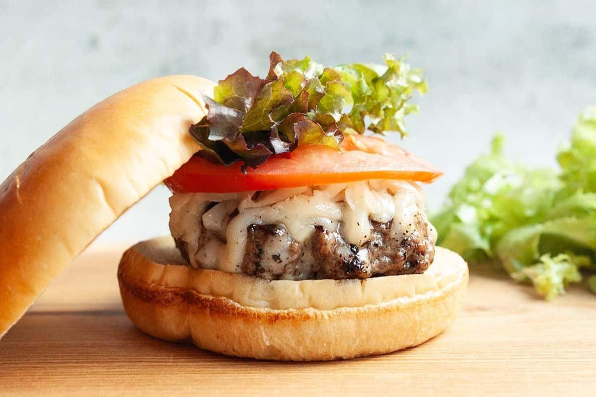 Open hamburger with melted cheese, tomato, and lettuce on a wooden surface, with lettuce in the background.