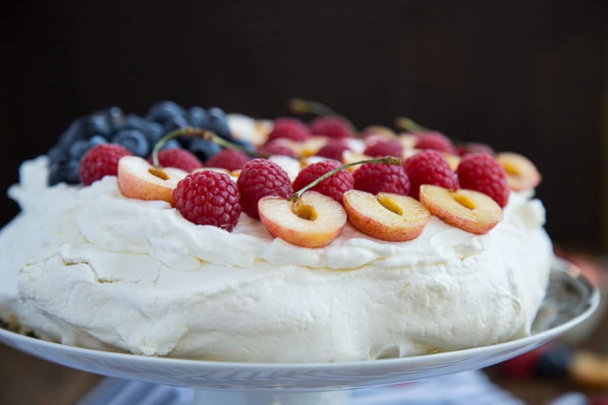 A pavlova topped with whipped cream, blueberries, raspberries, and halved cherries on a cake stand.