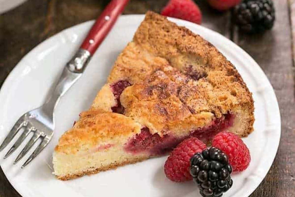 A slice of berry-filled cake on a white plate with a fork, raspberries, and a blackberry.