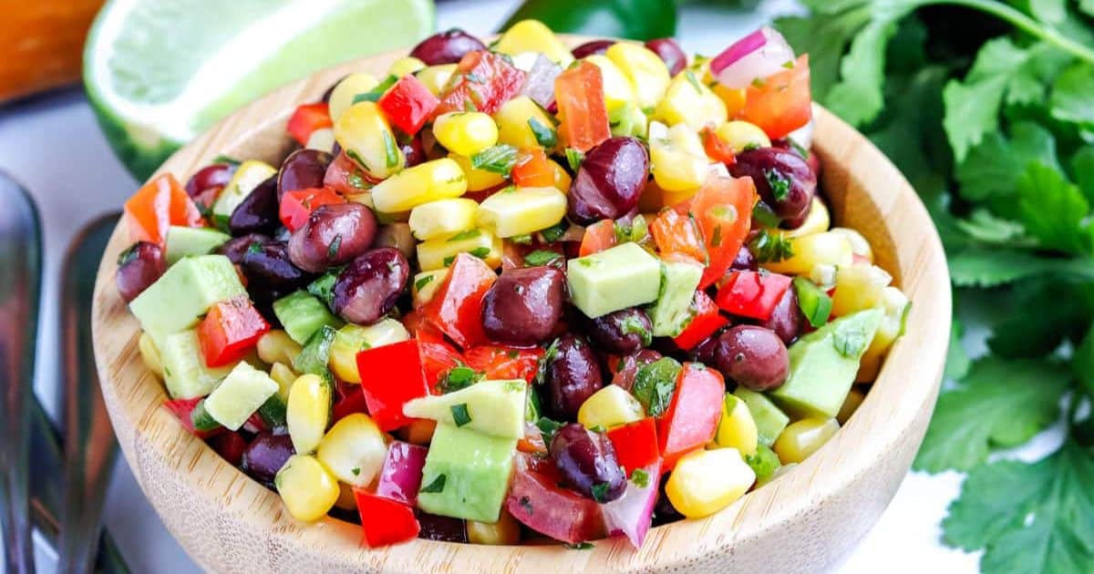 A wooden bowl filled with black bean, corn, avocado, tomato, and pepper salad with cilantro.