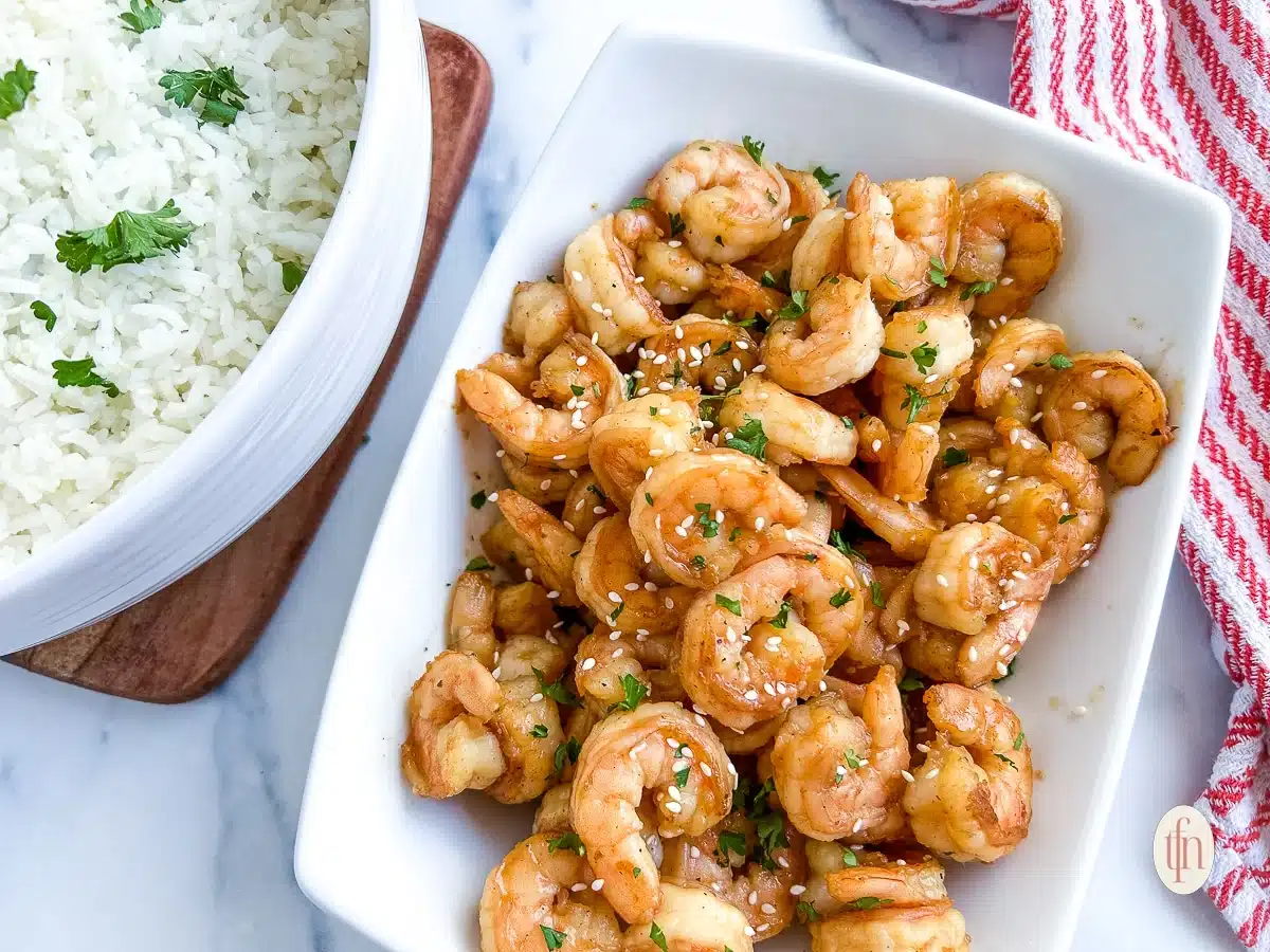 A white dish of glazed shrimp garnished with herbs and sesame seeds next to a bowl of rice.