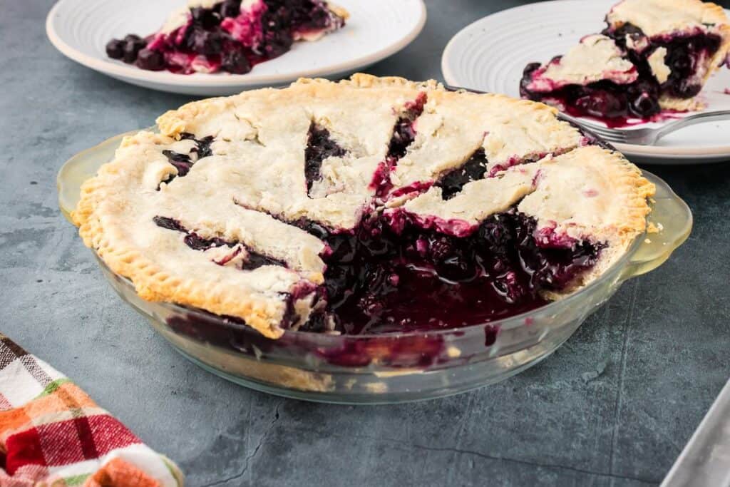 A sliced blueberry pie in a glass dish, with pieces served on white plates in the background.