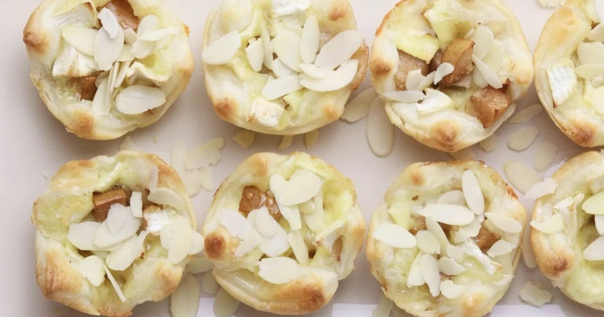 Mini pastry tarts filled with cheese, diced fruit, and topped with sliced almonds on a white surface.